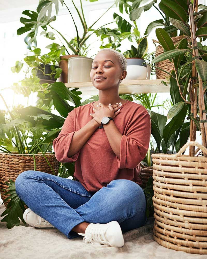 Meditation, Pflanze und schwarze Frau entspannen sich zu Hause für Wohlbefinden, Zen-Denkweise und ruhige Energie. Natur, Glück und das Gesicht einer weiblichen Person meditieren auf dem Boden mit umweltfreundlichen Farnen, Blättern und Zimmerpflanzen